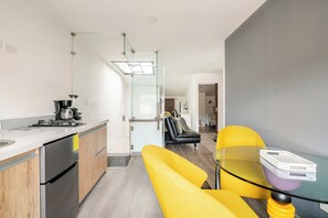 Une cuisine moderne équipée d’une machine à café, d’un réfrigérateur et d’une porte vitrée donnant sur un espace de vie avec un canapé et une table en verre.