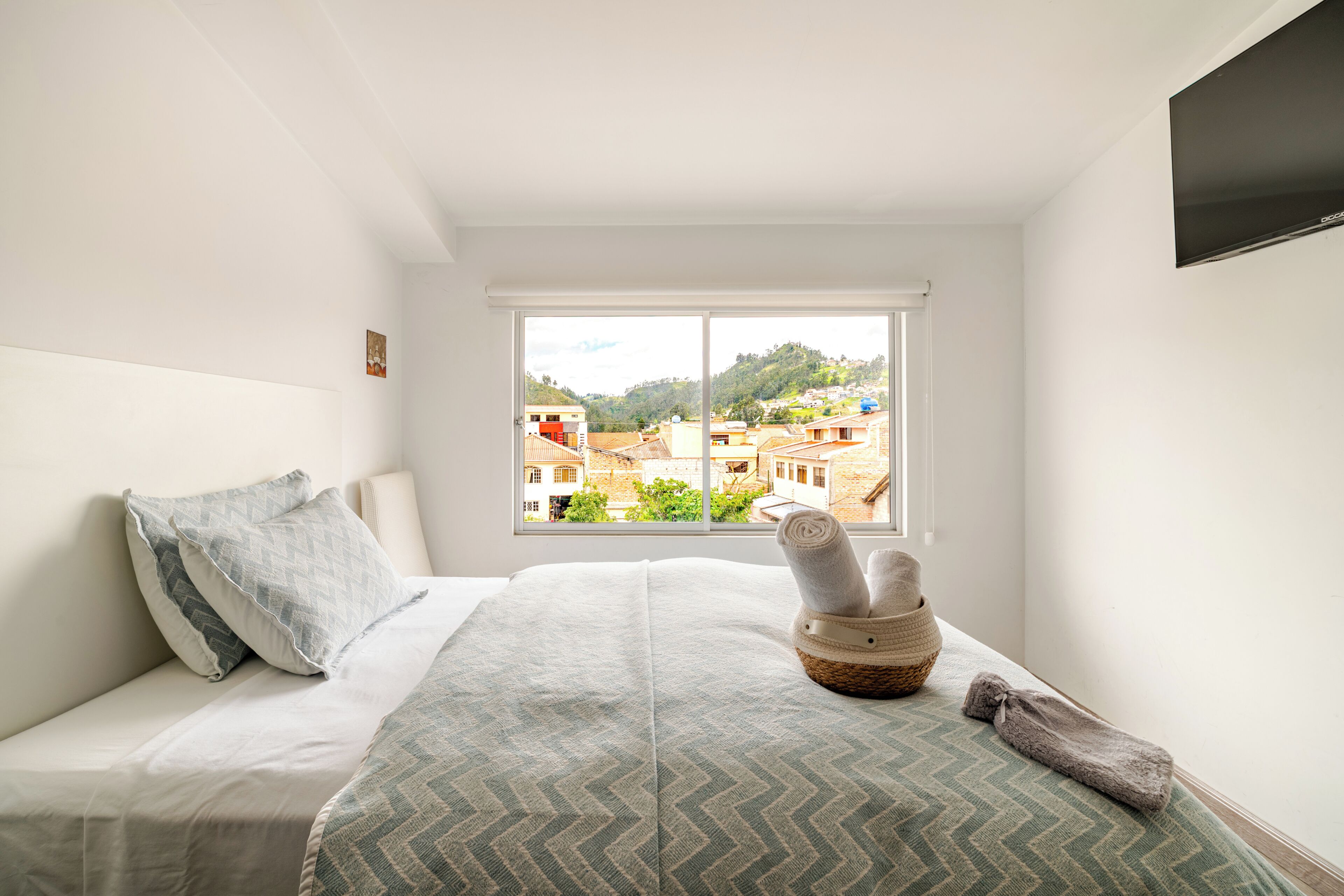 A bedroom with a bed, pillows, a window with a view of buildings, and a TV on the wall.