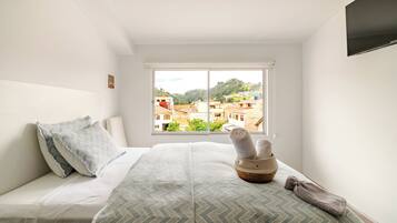 A bedroom with a bed, pillows, a window with a view of buildings, and a TV on the wall.