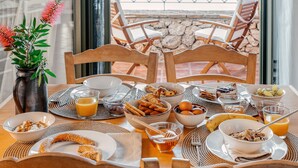 Tavola imbandita per la colazione con cereali, frutta e pasticceria, affacciata su un muro in pietra e sedie all'aperto.