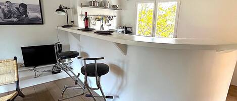 Una cocina moderna con una isla curva, taburetes de bar, un televisor y un ventanal con follaje amarillo.