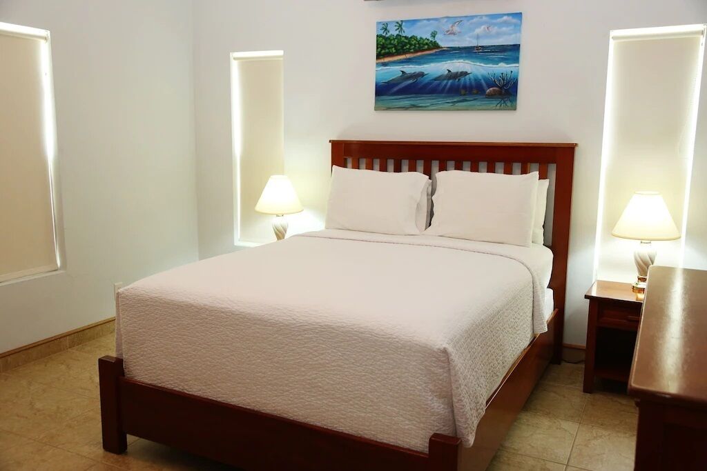 Una cama bien tendida con un cabecero de madera, dos lámparas de noche y un cuadro de una escena tropical sobre ella.