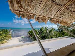 Uitzicht op een strand met een balkon, rieten dak, palmbomen en een helderblauwe lucht.