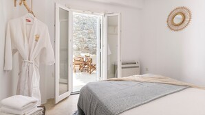 Quarto com cama, roupão pendurado, pequena mesa com cadeiras e vista para área externa através de uma porta de vidro.
