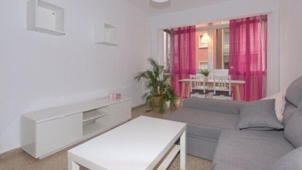 Ein Wohnzimmer mit grauem Sofa, weißem Couchtisch und einem weißen TV-Schrank. Es gibt einen Essbereich mit weißem Tisch und Stühlen, und eine Pflanze in der Ecke.
