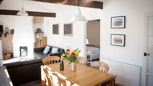 A dining area with a wooden table, chairs, and a vase of flowers. There are two pendant lights above the table. - Contemporary cottage in lovely Devon village. (Musbury)