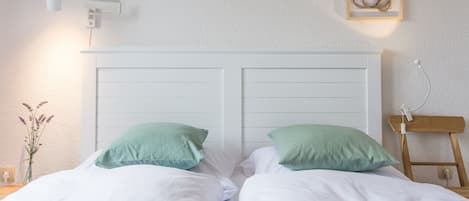 A bedroom with a white headboard, two green pillows, a bedside table with a lamp, and framed artwork on the wall.