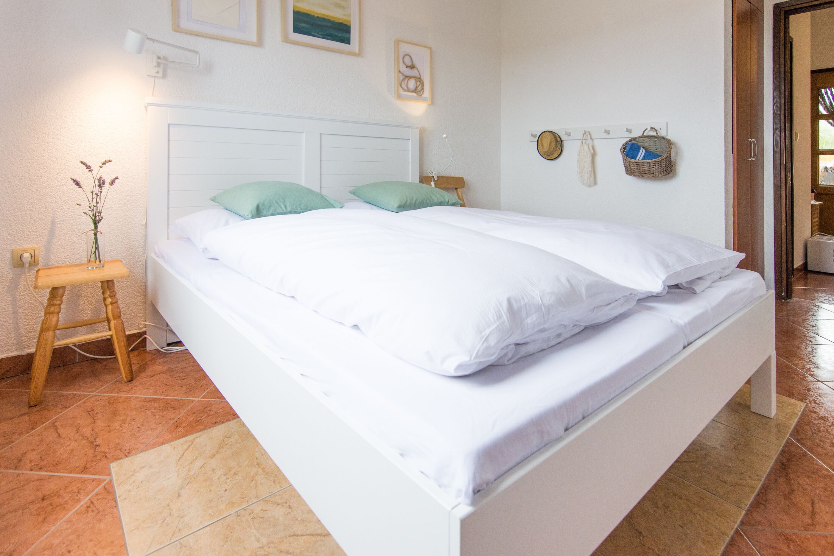 Un letto rifatto con lenzuola bianche e due cuscini verdi, un comodino con un vaso di fiori e una lampada fissata al muro.