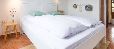 A neatly made bed with white linens and two green pillows, a bedside table with a vase of flowers, and a wall-mounted lamp.