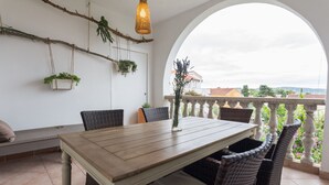 Ein Essbereich mit Holztisch und Korbstühlen, einer Bank, hängenden Pflanzen und Blick durch ein Rundbogenfenster.