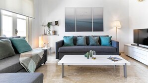 Una sala de estar moderna con un sofá gris, cojines color turquesa, una mesa de centro blanca y un televisor grande sobre un mueble blanco.