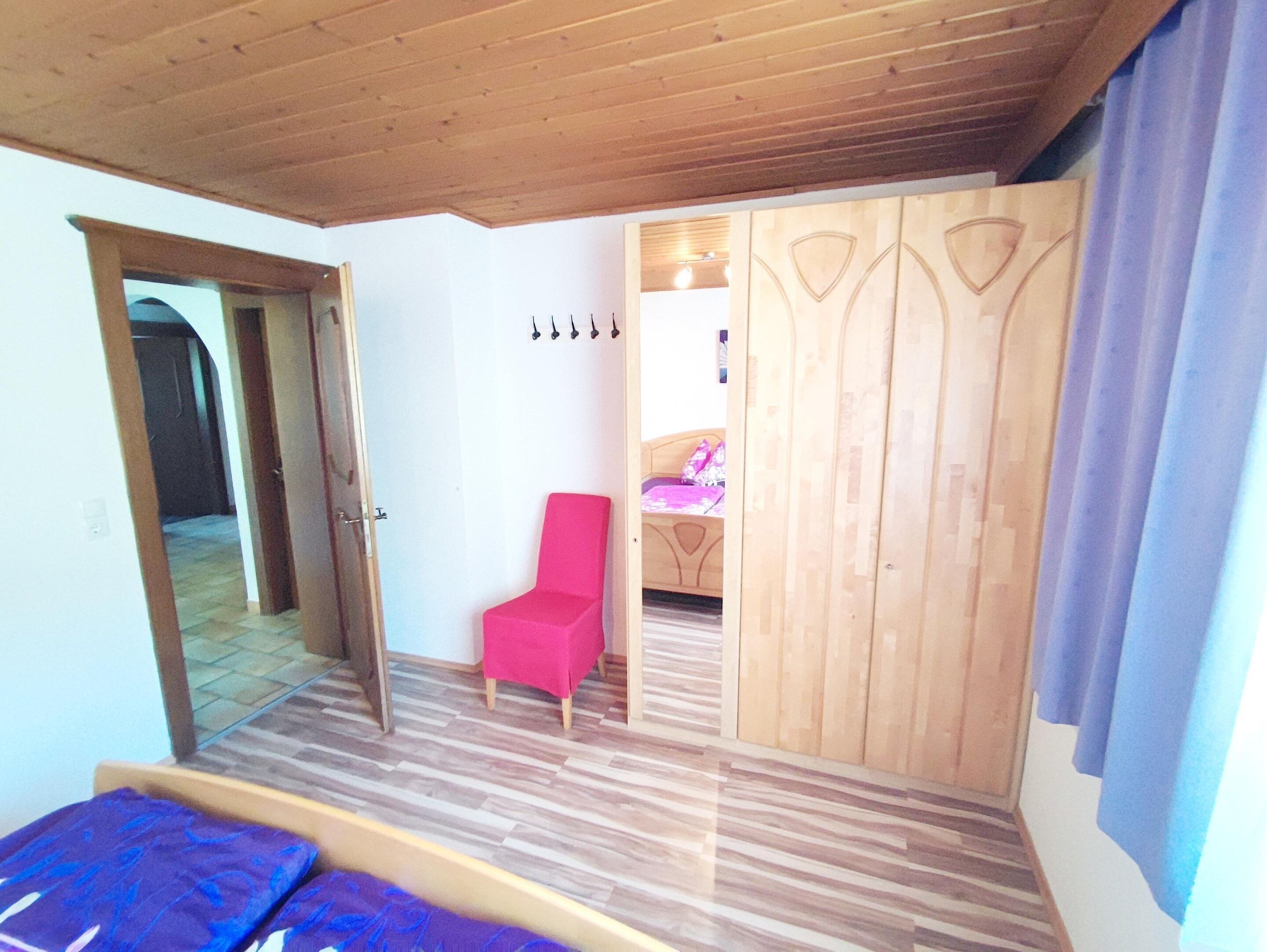 A room with a wooden ceiling, a bed with a blue bedspread, a red chair, and a wooden wardrobe with a mirror.