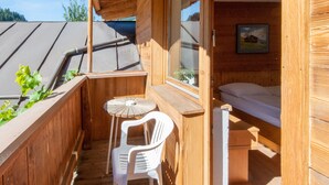 Ein hölzerner Balkon mit einem kleinen Tisch und einem weißen Plastikstuhl, ein Fenster mit Blick zum Himmel sowie ein Bett mit Matratze und Kissen.