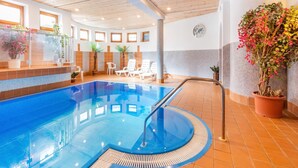 Ein beheiztes Hallenbad mit glasklarer Wasseroberfläche, umgeben von Liegestühlen und Topfpflanzen.