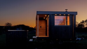 Une petite cabane en bois, dotée d’une seule porte et d’une fenêtre, située dans un champ dégagé, avec un coucher de soleil en arrière-plan.