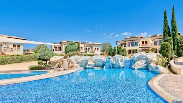 A swimming pool with a rocky waterfall feature, surrounded by well-maintained landscaping and modern buildings. - Villa TA01 Ouranos - Three Bedroom Villa, Sleeps 6 (Kouklia)