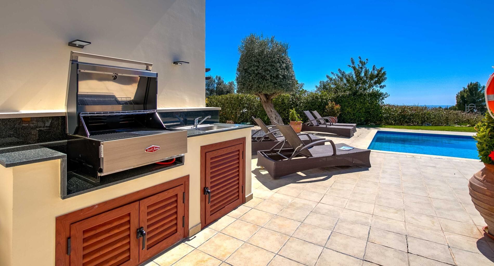 Une cuisine extérieure équipée d’un barbecue et d’un évier, entourée de fauteuils et d’une piscine.