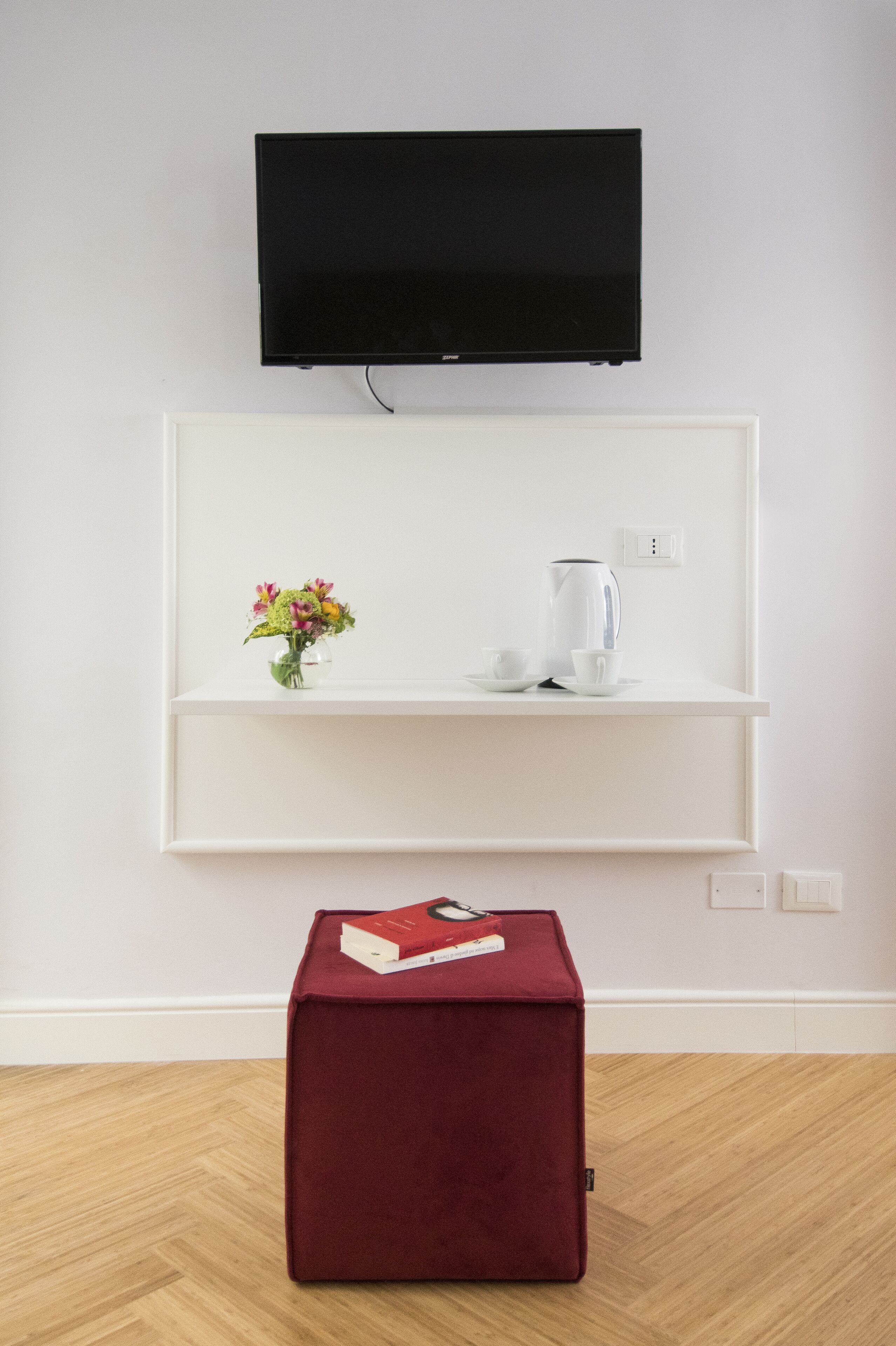 A room with a mounted TV, a white shelf with a vase of flowers, a white kettle, and a red ottoman with a book.