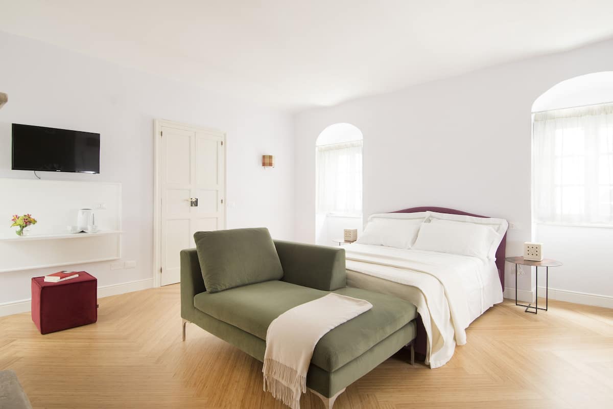 A modern bedroom with a green sofa, a bed with white bedding, a TV on the wall, and a small table with a lamp.