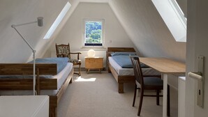 Une petite chambre mansardée, bien éclairée, avec deux lits en bois, un bureau et une chaise. Une fenêtre laisse entrer la lumière naturelle et offre une vue sur la verdure environnante.