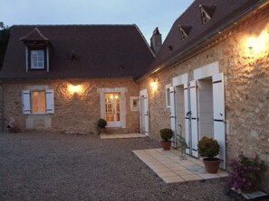Un bâtiment en pierre avec une allée en gravier, une petite entrée dotée d’une porte et un pot de fleurs placé près de l’entrée.