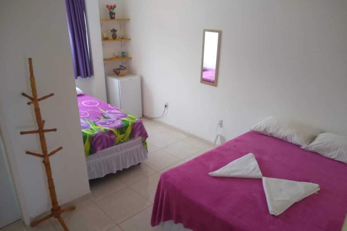 Un dormitorio con dos camas, una con cubrecama morado y almohadas blancas, y otra con cubrecama colorido. Se observa un perchero de madera y un pequeño armario blanco con un espejo en la pared.
