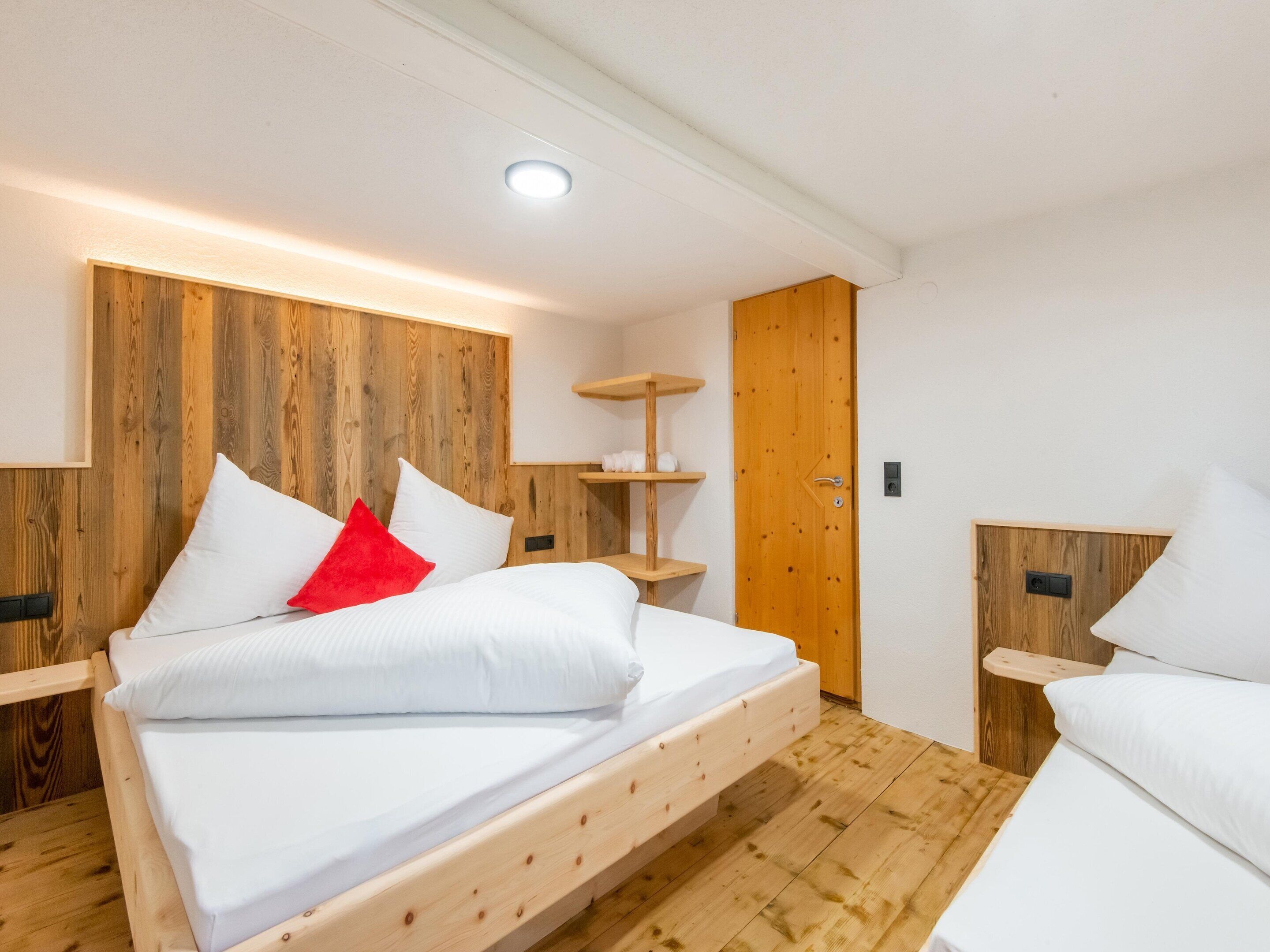 Quarto moderno com cabeceira de madeira, cama com roupa de cama branca e um travesseiro vermelho, chão de madeira e porta de madeira.