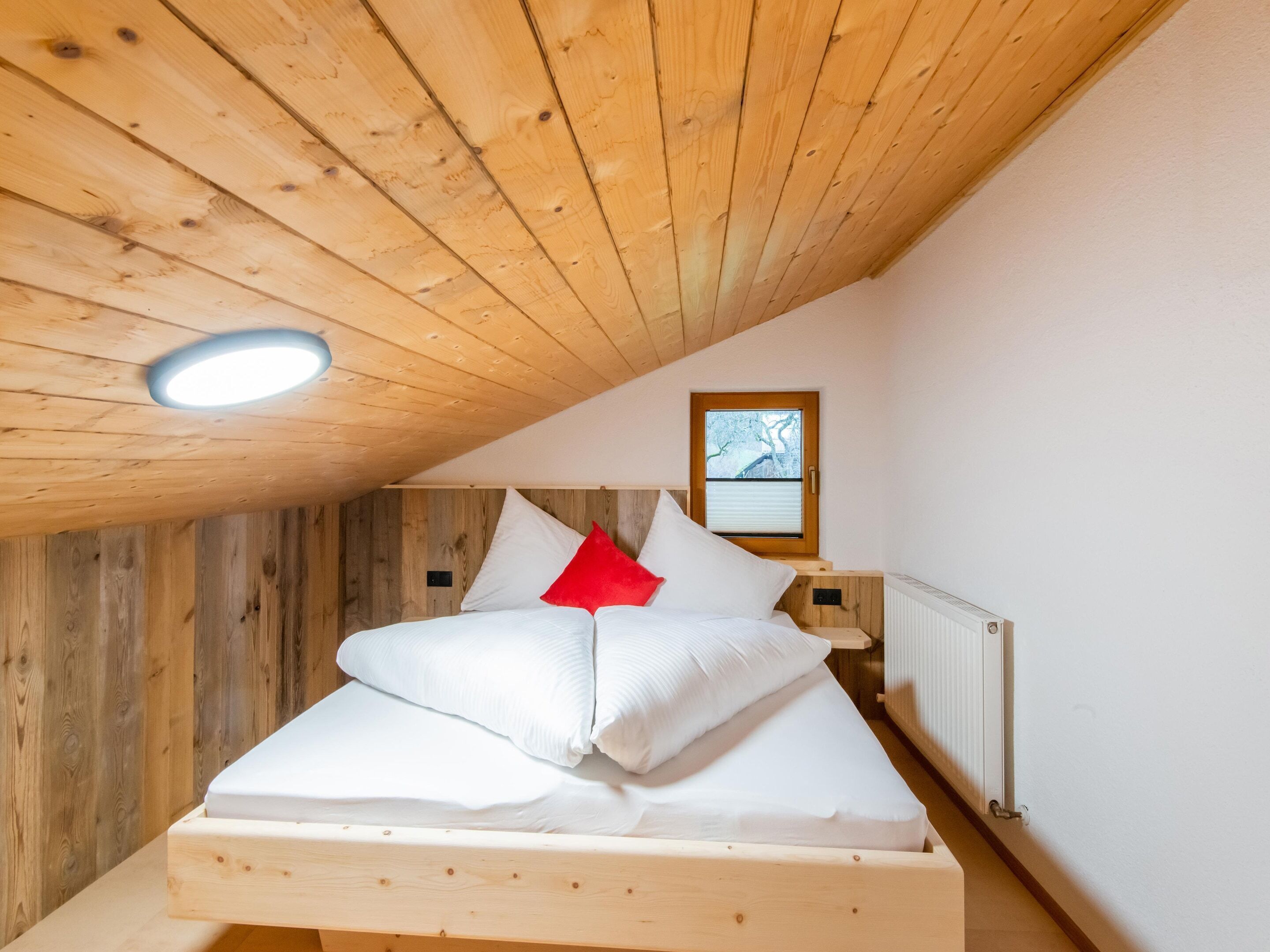 Un dormitorio acogedor con techo inclinado, paredes de madera, una cama con ropa de cama blanca y almohada roja, una ventana pequeña con vistas, y una mesita de noche con lámpara.