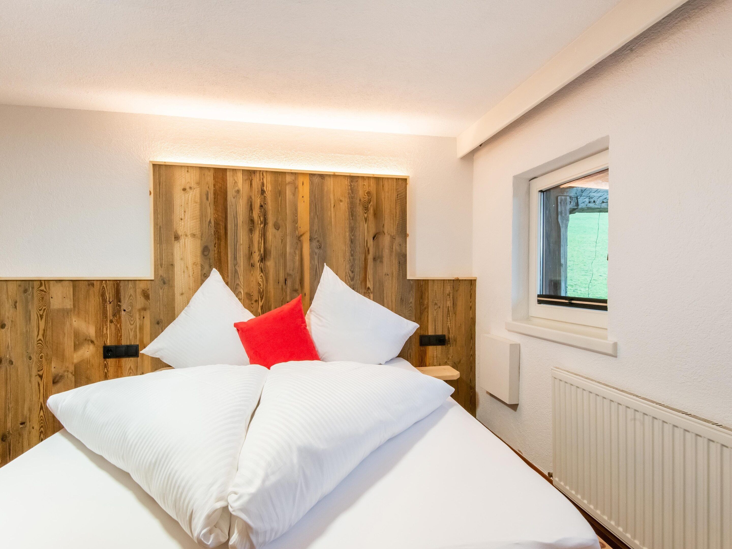 Un lit bien fait, avec des oreillers blancs et rouges, une tête de lit en bois et une fenêtre donnant sur un paysage verdoyant.