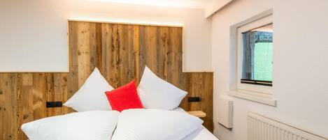 Un lit bien fait, avec des oreillers blancs et rouges, une tête de lit en bois et une fenêtre donnant sur un paysage verdoyant.