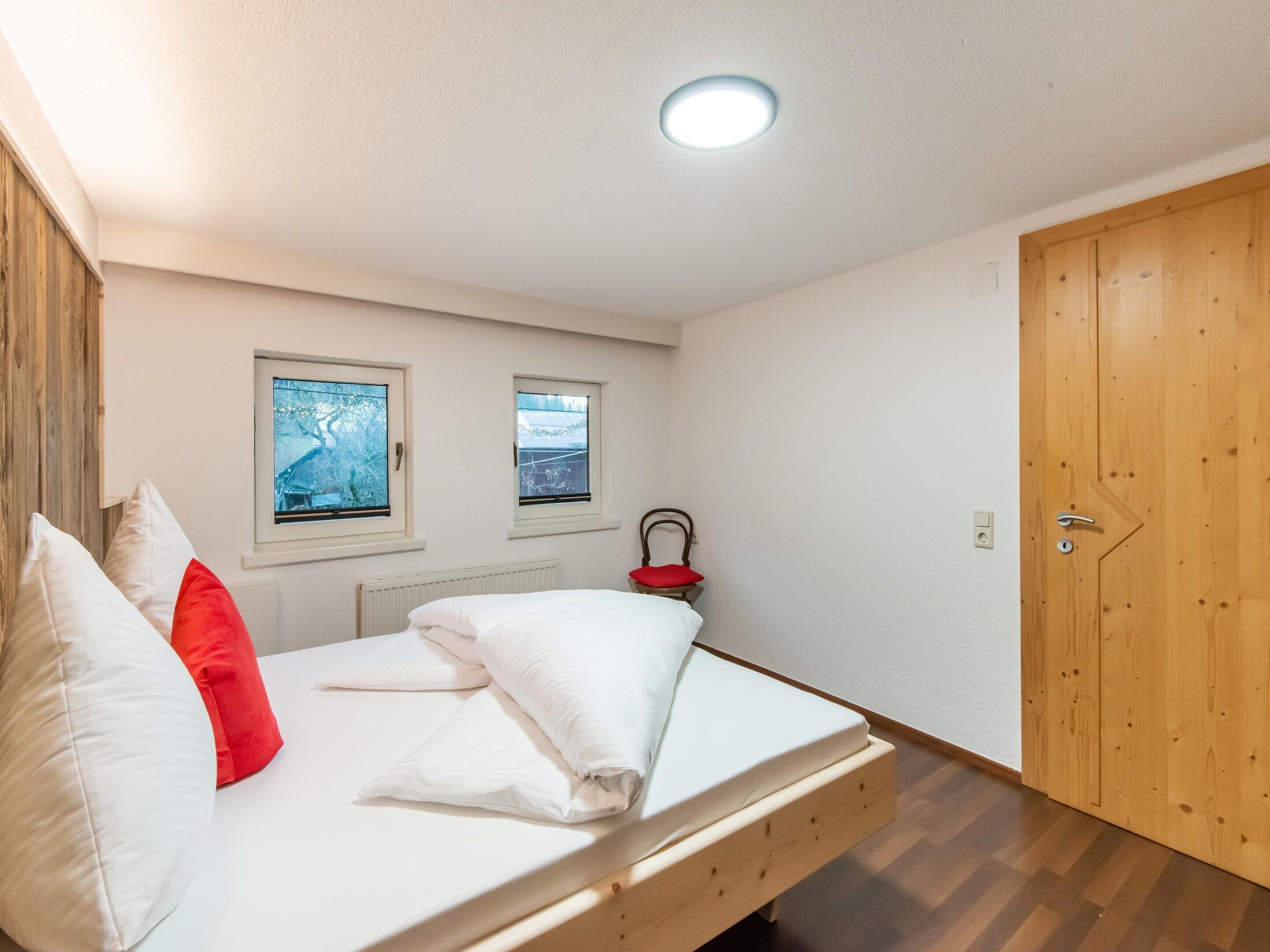 Une chambre à coucher avec un lit, deux fenêtres, un radiateur, une porte en bois et un luminaire au plafond.
