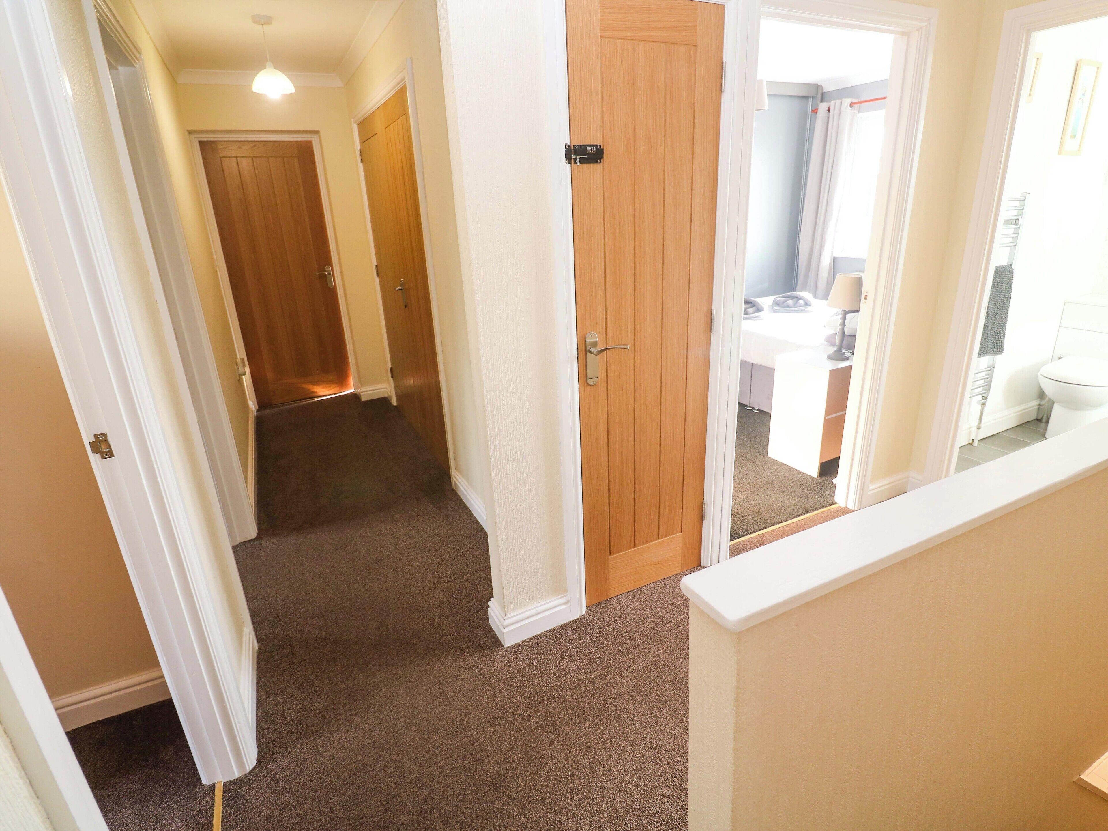 A hallway with carpeted floors leading to a wooden door, a white wall, and a glimpse of a bathroom with a toilet and sink.
