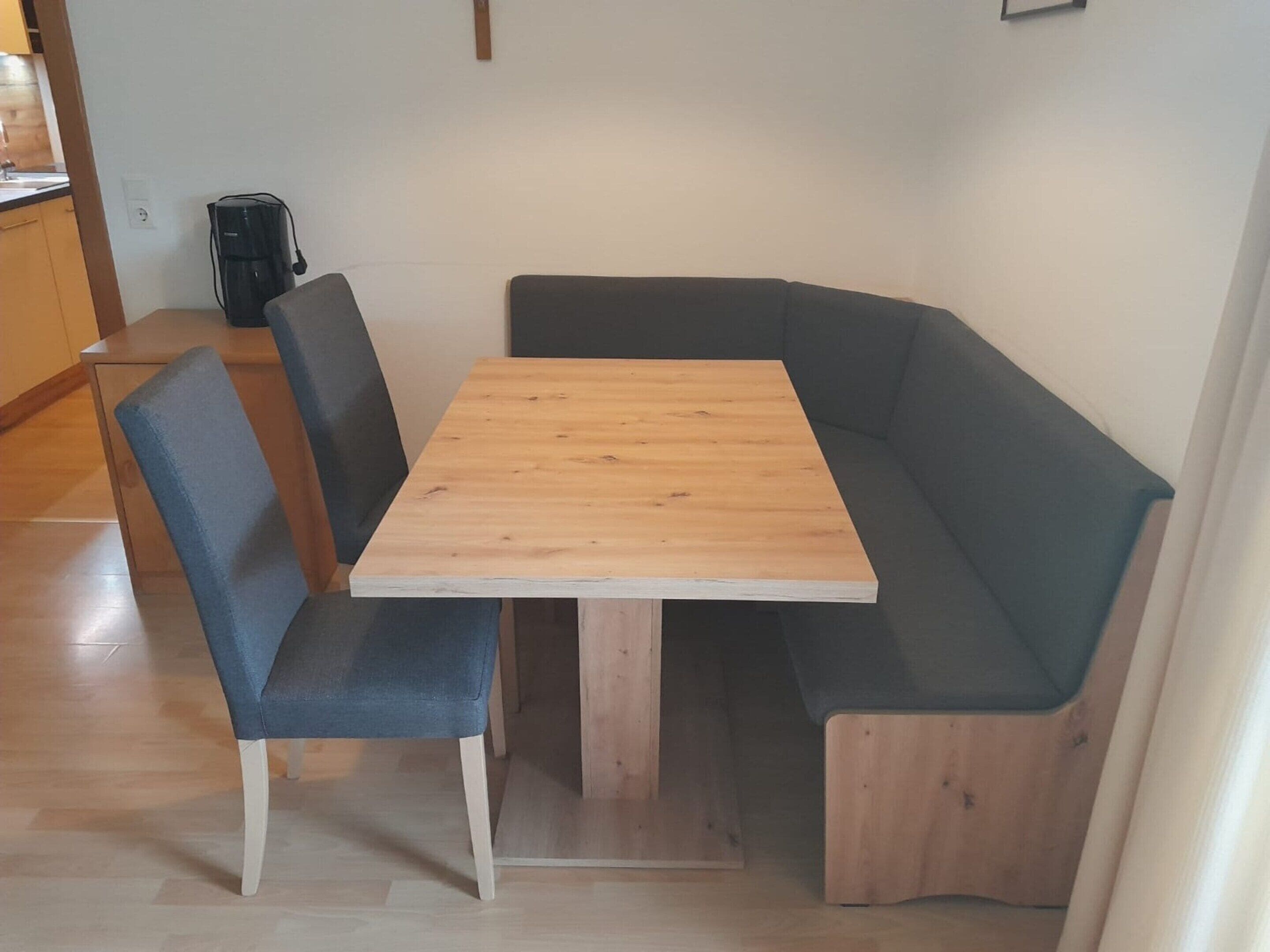 Ein Essbereich mit einem Holztisch, zwei blauen Stühlen, einer schwarzen Kaffeemaschine und einem Holzschrank.