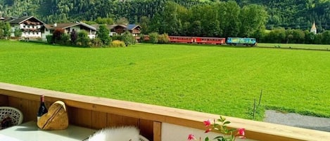 A terrace with a view of a green field and a red train in the distance.