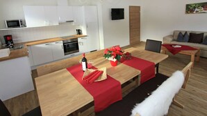 Moderni keittiö, jossa on valkoiset kaapit, mikroaaltouuni ja kahvinkeitin. Ruokailutilassa on puinen pöytä, punaiset pöytäliinat ja kattokruunu.