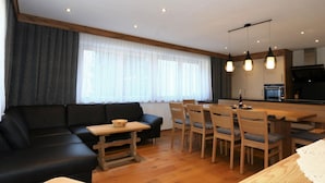 Una sala de estar moderna con un sofá negro, una mesa de comedor de madera y sillas. Se observa una cocina al fondo con estufa y armarios.
