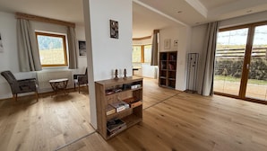 A spacious living room with wooden flooring, a built-in bookshelf, a dining area with a table and chairs, and large windows offering a view of the outdoors. - Family Apartment in Neustift With Sauna and Garden (Neustift Im Stubaital)