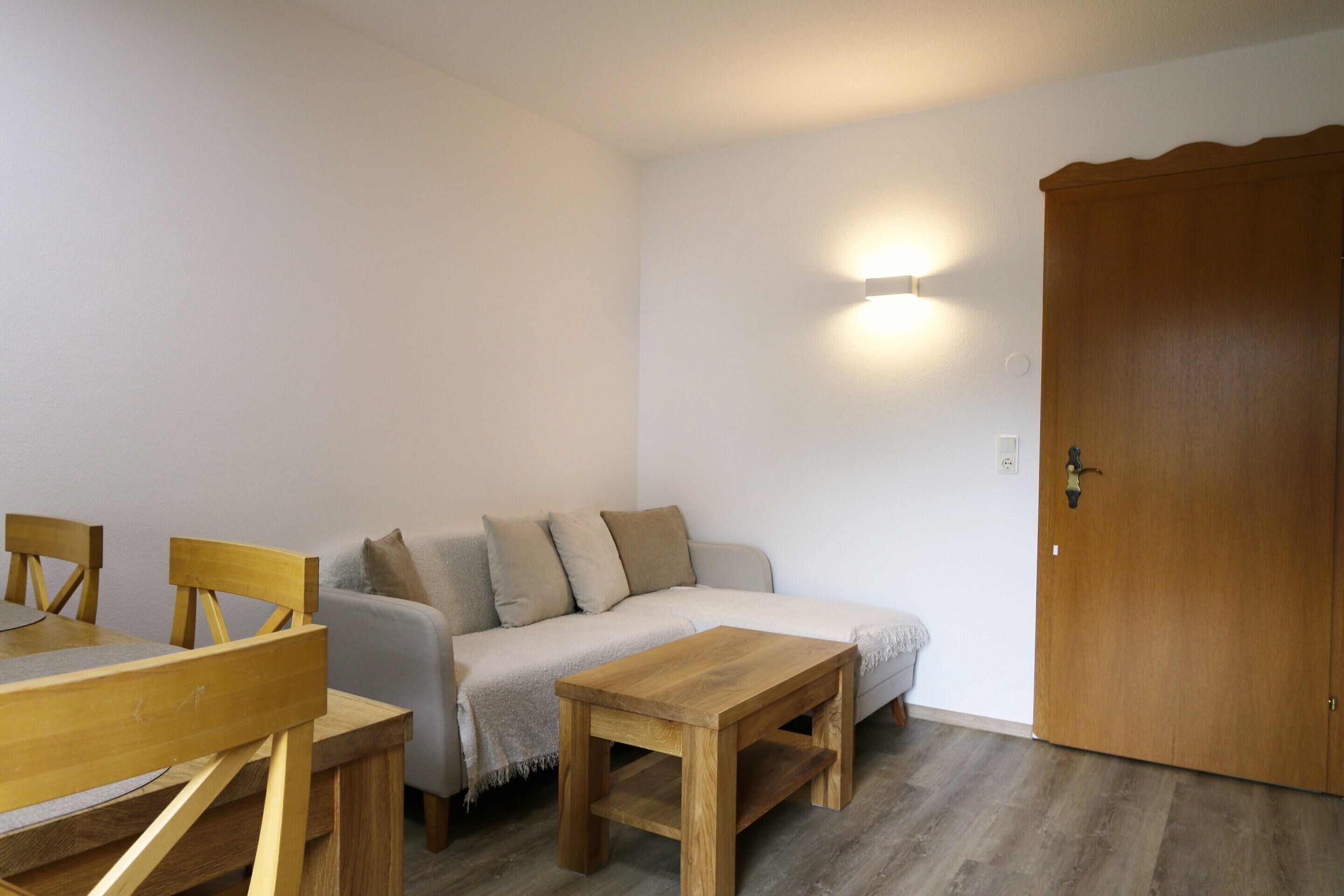 Ein Zimmer mit einem Holzesstisch, zwei Stühlen, einem grauen Sofa mit Kissen, einem Holzhocker und einer Holztür.