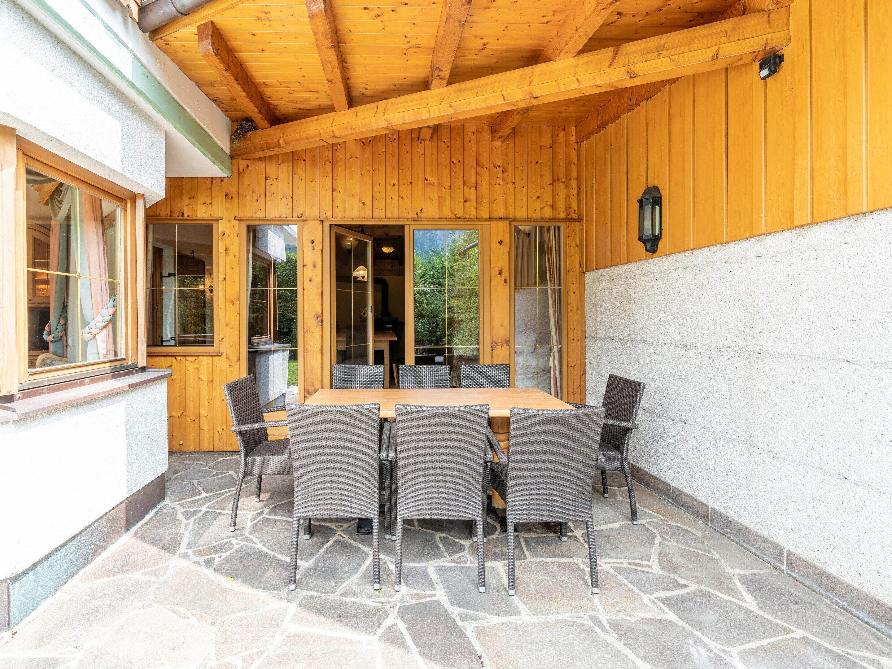 Eine Holzterrasse mit Steinplatten, ein Holztisch mit Stühlen und eine Glastür zum Garten.
