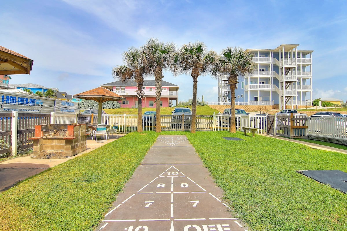A shuffleboard court with a clear path, surrounded by a fenced area, palm trees, and residential buildings in the background.