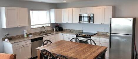 A modern kitchen with white cabinets, a stainless steel refrigerator, a built-in oven, and a wooden dining table with four chairs.