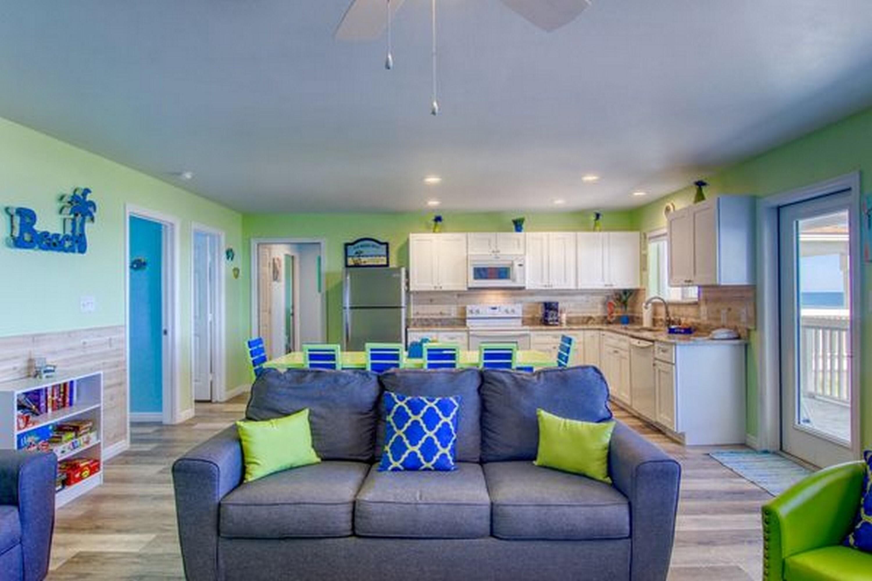 A living room with a gray sofa, a dining area with blue chairs, and a kitchen with white cabinets.