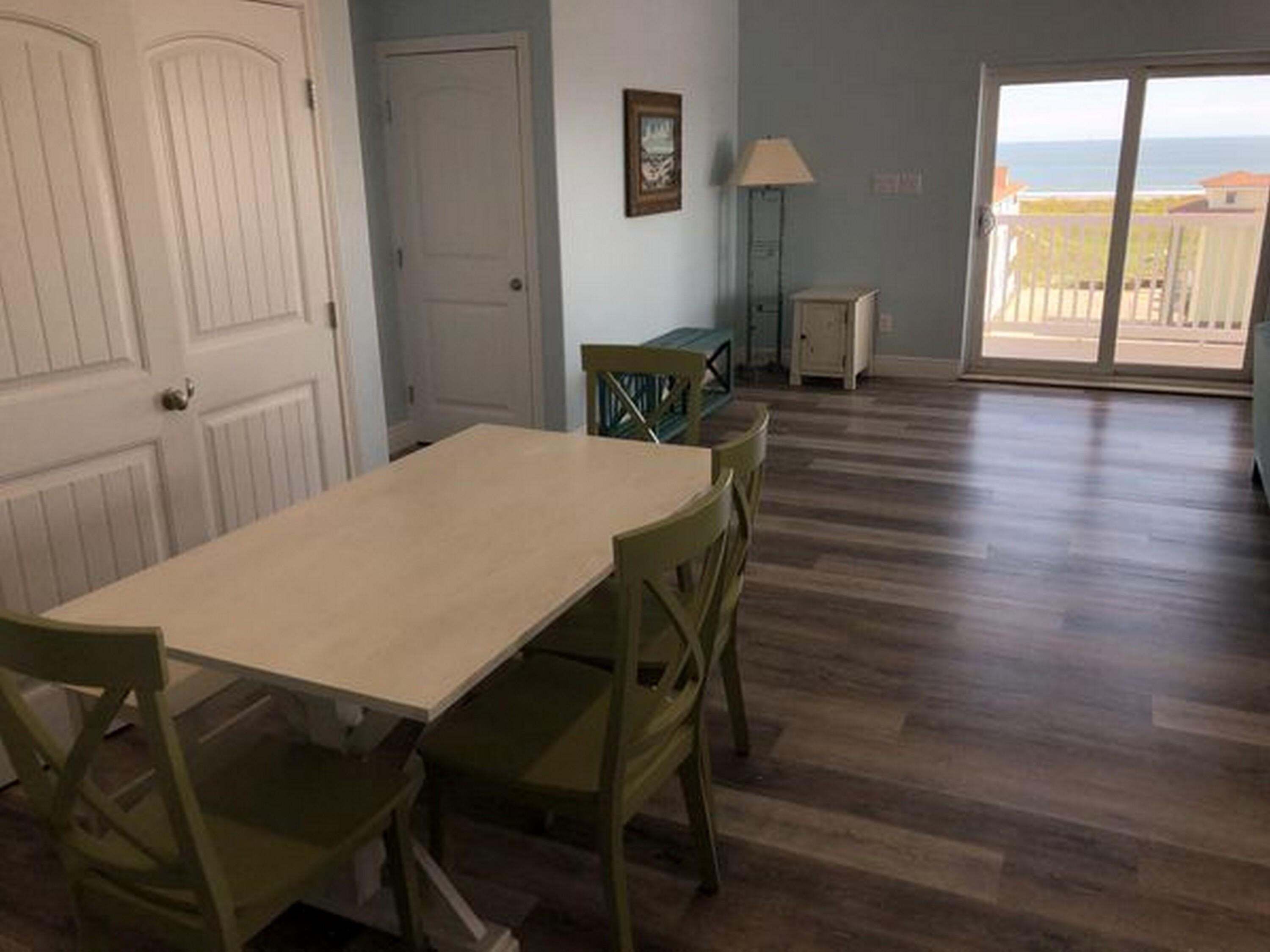 Een kamer met een eettafel, stoelen, een lamp, een klein kastje en uitzicht op de oceaan door een schuifglazen deur.