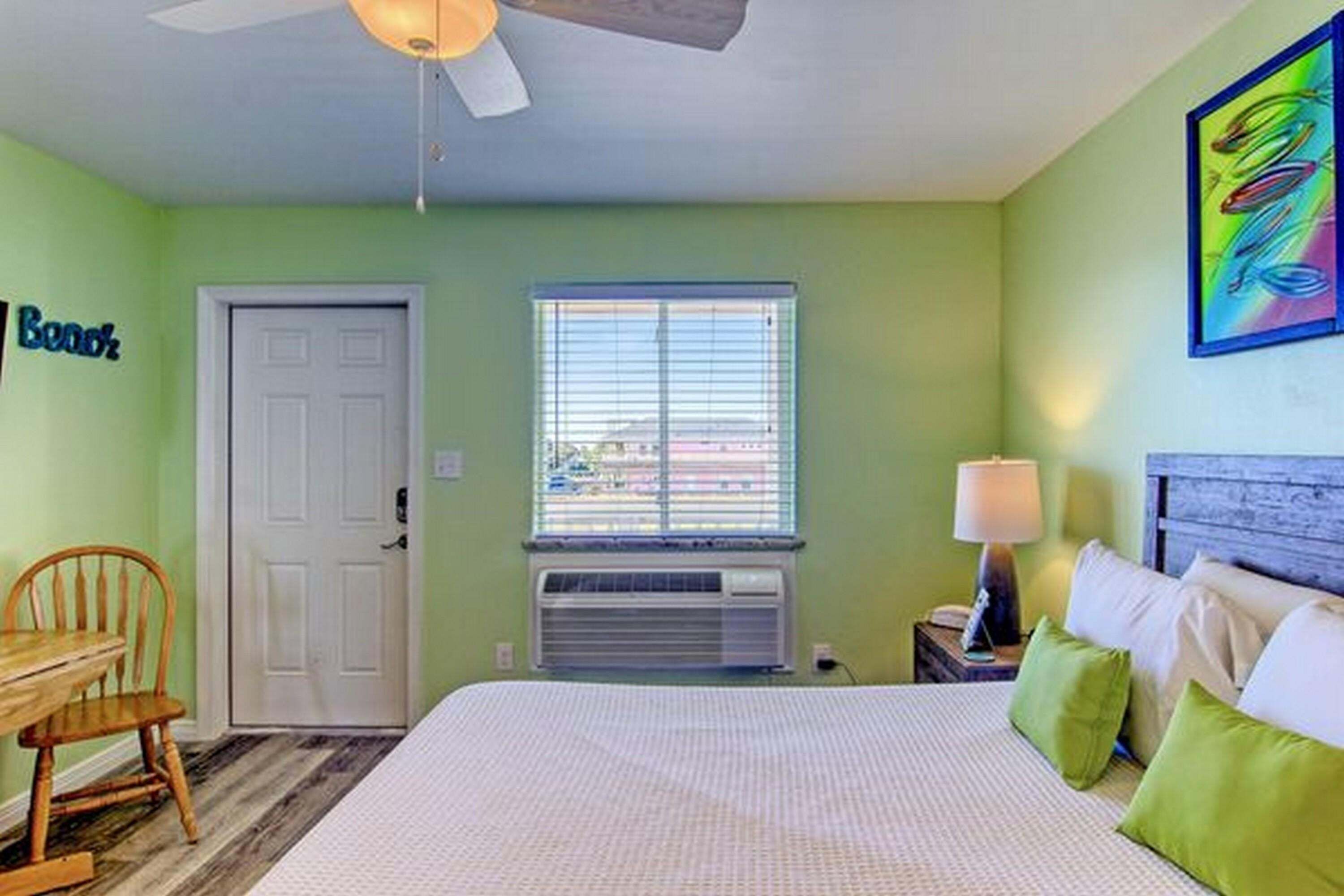 A bedroom with a bed, a desk, a chair, a window with blinds, a ceiling fan, and a wall-mounted air conditioner.