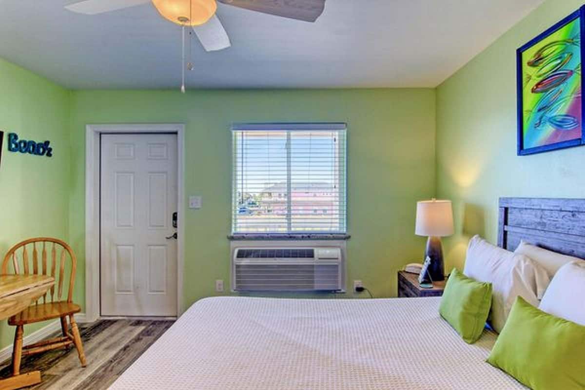 A bedroom with a bed, a desk, a chair, a window with blinds, a ceiling fan, and a wall-mounted air conditioner.
