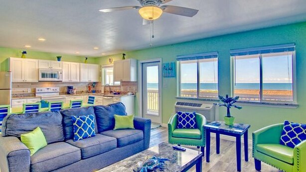 A spacious living area with a gray sofa, green armchairs, and a coffee table. A ceiling fan and a view of the ocean are visible. - Beachgate CondoSuites and Oceanfront Resort (Port Aransas)