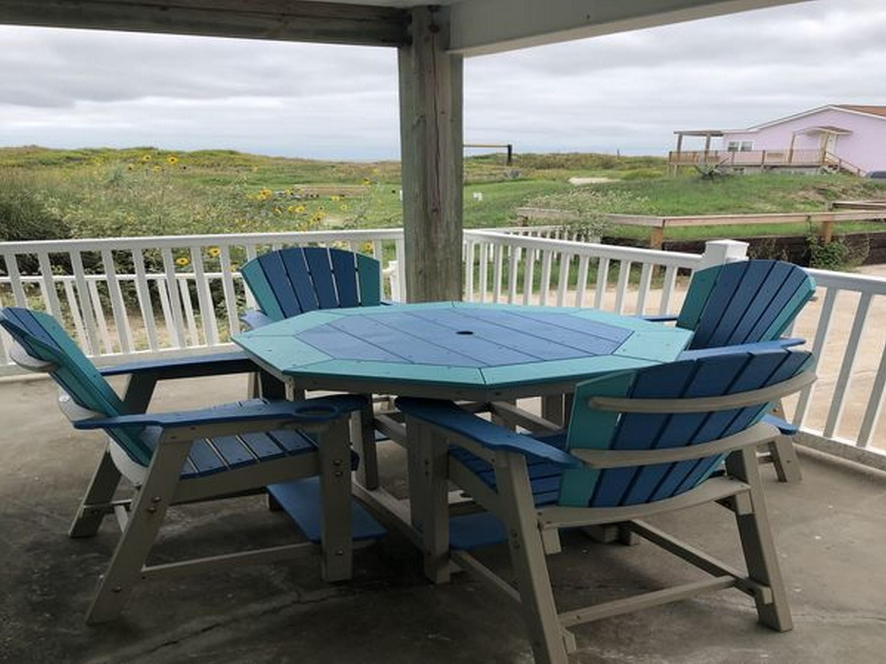 A covered outdoor seating area with a table and chairs, a white railing, and a view of a grassy landscape with a building in the background.
