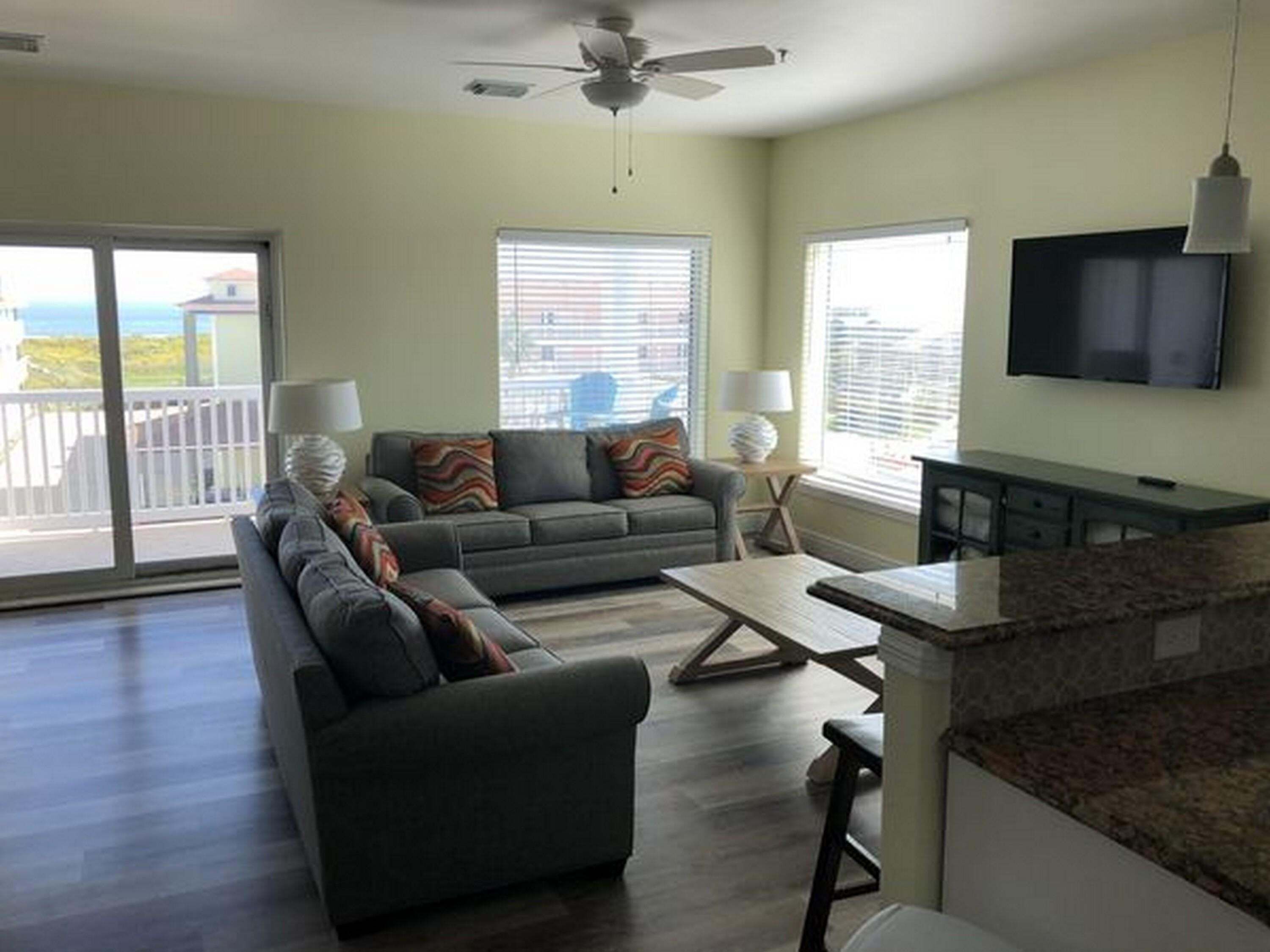 A living room with a sofa, a coffee table, and a TV. There is a balcony with a view.
