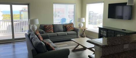 A living room with a sofa, a coffee table, and a TV. There is a balcony with a view.