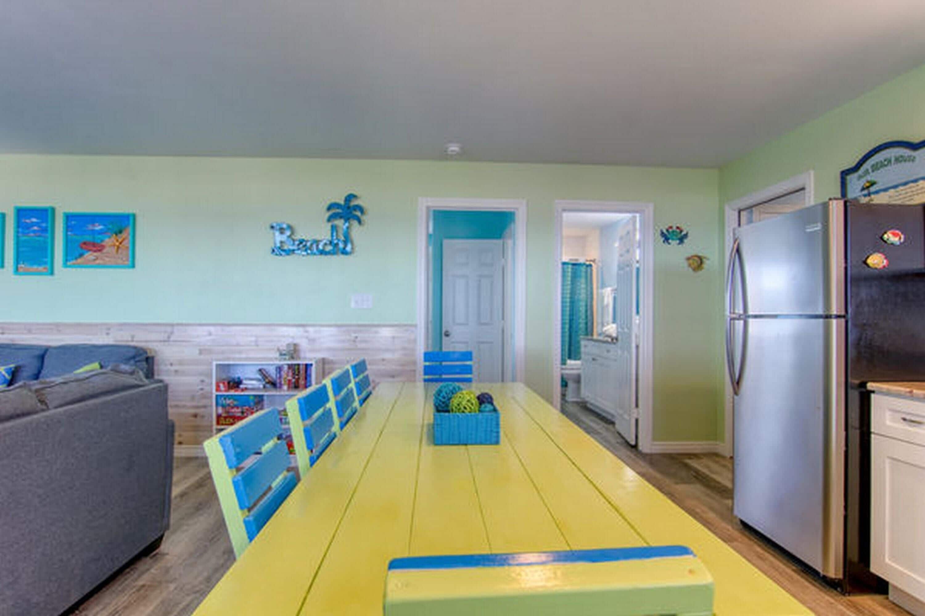 A bright kitchen with a yellow table, blue chairs, a grey sofa, and a fridge.